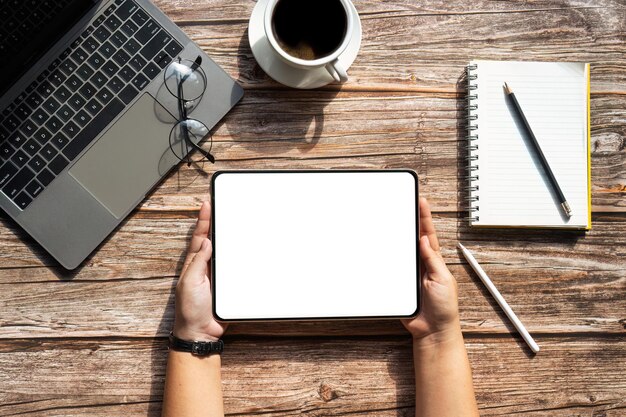 Top view tablet that someone is holding with blank screen mock up digital tablet on workspace office desk with cup of coffee notebook and laptop