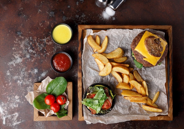 Top view of take away meal at old rusty background, copy space.\
wooden tray with black bun cheeseburger and baked potato wedges,\
spilled salt, dipping sauces set and cherry tomatoes, fast food\
concept