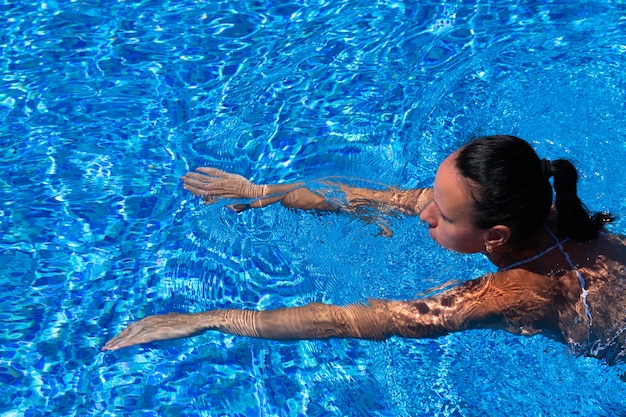 Top view of a tanned girl, female, model in a red swimsuit, swimming in the blue water of the pool.