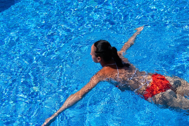 Top view of a tanned girl, female, model in a red swimsuit, swimming in the blue water of the pool.