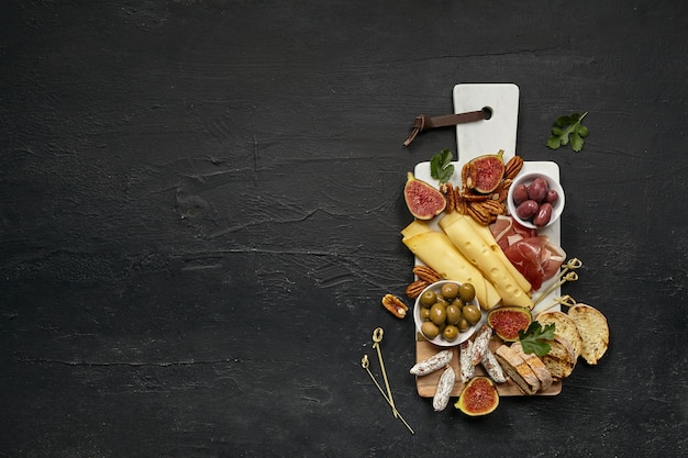 Photo top view of tasty cheese plate with fruit, grape, nuts, olives and toasted bread on a wooden kitchen plate on the black stone background, top view, copy space. gourmet food and drink.