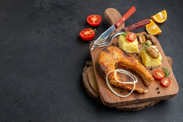 Top view of tasty fried fish and mushrooms tomatoes greens on cutting board cutlery set pepper on black surface