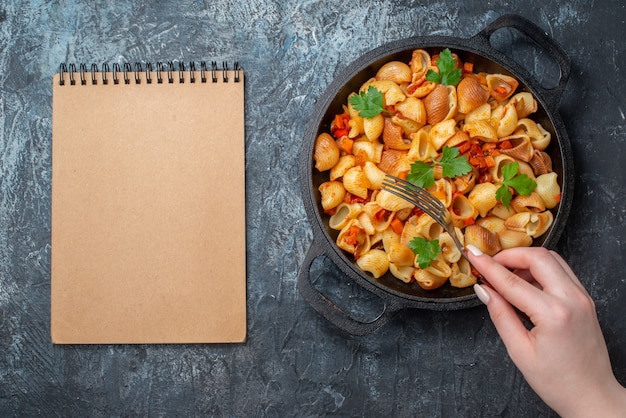 top view tasty pasta in frying pan spaghetti rigatoni spirals cherry tomatoes fork in woman hand notebook on grey background
