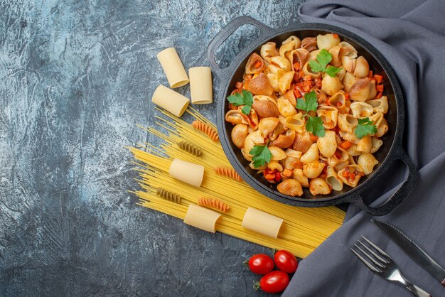 Top view tasty pasta in frying pan spaghetti rigatoni spirals cherry tomatoes knife and fork on grey background copy place