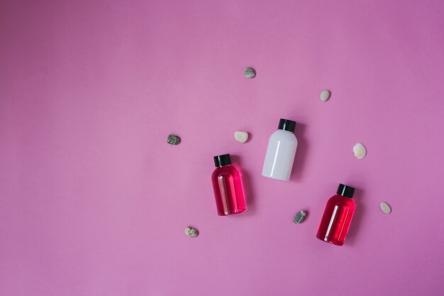 Vista dall'alto di tre piccole bottiglie di bianco e cremisi e prodotti per la cura del corpo e dei capelli, ciottoli di mare sulla cima di uno sfondo rosa. cosmetici per i turisti