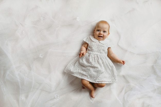 top view of tiny baby girl with sincere smile, lying on back on white bed sheet, dressed in white dress and looking straight