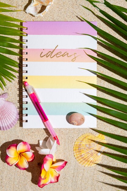 Top view of Travellers notebook with pen on sand with palm tree, flowers, sea shells.