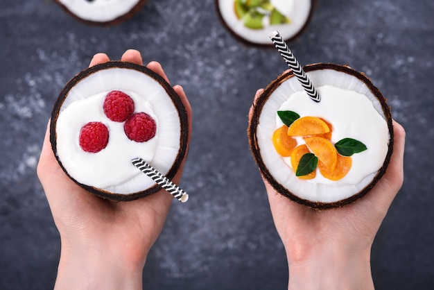 Vista dall'alto due dessert con crema e frutti di bosco in noce di cocco sulle mani su sfondo grigio, concetto di ciotole di frullato