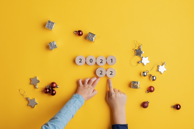 Top view of two kids changing 2021 sign made of wooden cut circles into a 2022 sign. Over yellow background with holiday decorations scattered around.