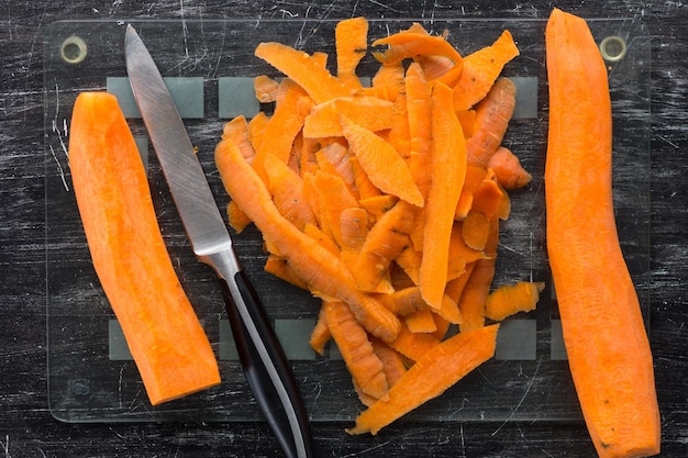 Top view of two peeled carrots with peeling and knife on glass cutting board on the black background