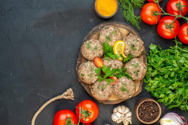 Top view of uncooked meatballs on a wooden cutting board and fresh vegetables green yellow ginger on dark background