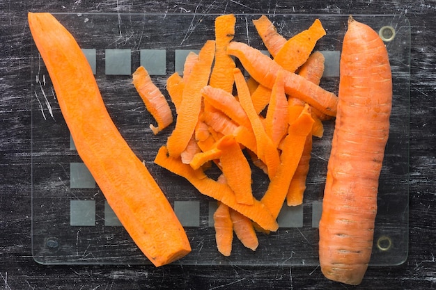 Top view of unpeeled carrot and peeled carrot with peeling on glass cutting board on the black background