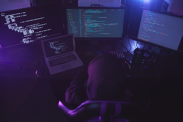 Vista dall'alto di un hacker di sicurezza informatica irriconoscibile che indossa il cappuccio mentre si lavora sul codice di programmazione in camera oscura, copia dello spazio