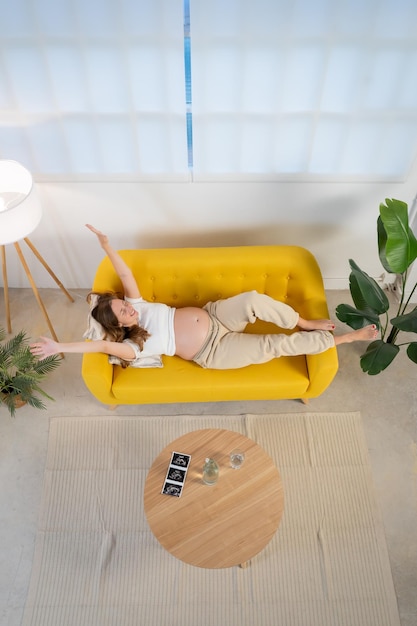 Top view van zwangere vrouw rust op gele bank in een gezellige woonkamer gelukkig met uitgestrekte armen