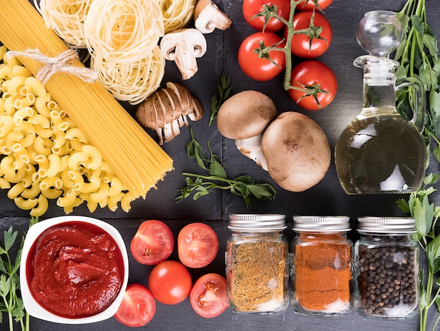 Top view variety of raw uncooked pasta next to a bowl with tomato souce, fresh tomatoes, mushrooms, greenery, different spices and a bottle with sunflower oil