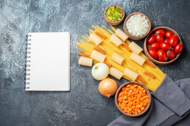 top view various pastas rigatoni spaghetti spirals chopped vegetables in bowls notepad on grey background