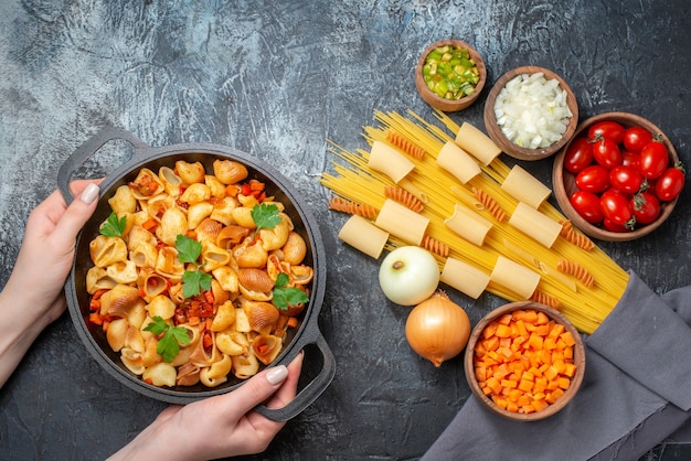 top view various pastas rigatoni spaghetti spirals chopped vegetables in bowls tasty pasta in frying pan in female hands on grey background