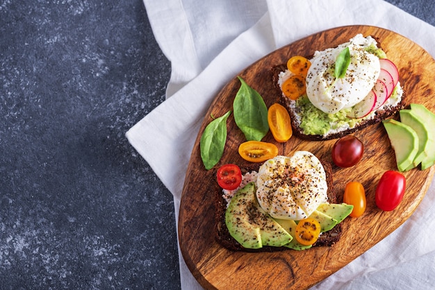 Top view vegetarian toast with poached eggs, cottage cheese, avocado, spinach, cherry tomatoes on wooden Board on gray background