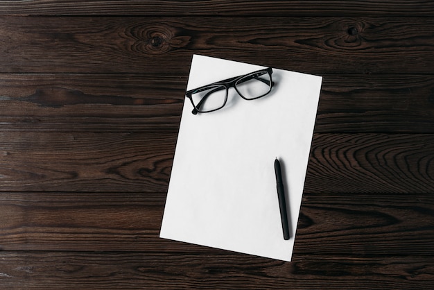 Top view of a white blank sheet of paper with a pen and glasses on a wooden table.