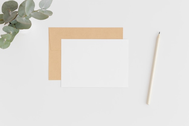 Top view of a white card mockup and envelope with a pen and eucalyptus on a white table