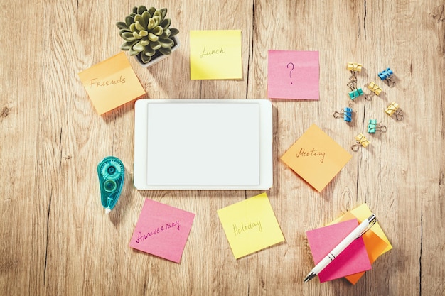 Top view of a white digital tablet with blank screen and many sticky papers with plans and ideas on wooden table.