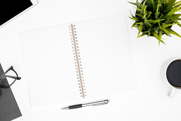 Photo top view of white office desk table with blank notebook with grid lines and supplies.