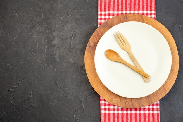 Top view white plate with wooden fork and spoon on wooden cutting board dark surface