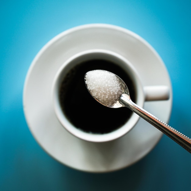 Top view of white sugar in aluminium spoon over black hot coffee cup on blue background