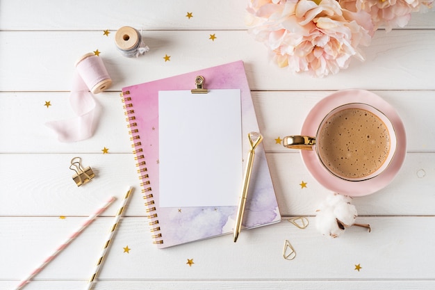 Top view of white working table  with blank paper notebook, cup of coffee. Flat lay peonies flowers, golden paper binder clips. Notepad and pen.