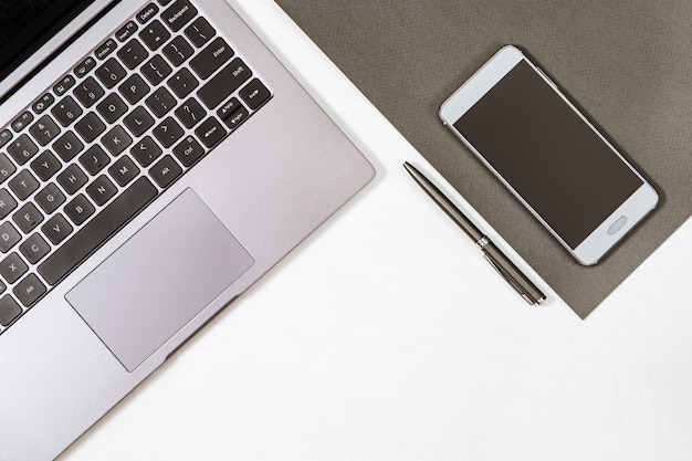 Vista superiore del tavolo di lavoro bianco con computer portatile, penna e smartphone grigi.