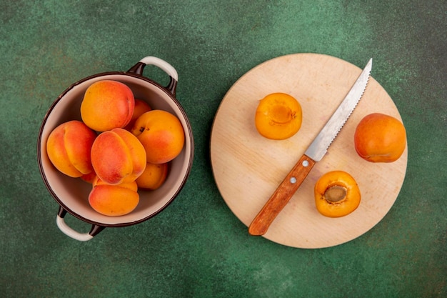 Photo top view of whole apricots in bowl and half cut one with knife on cutting board on green background