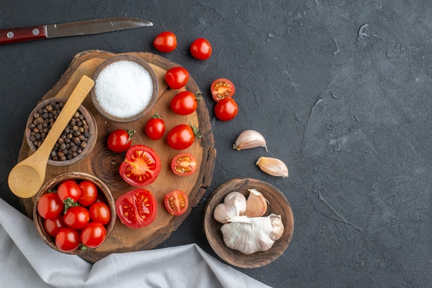 Top view of whole cut fresh tomatoes and spices on wooden board white towel garlics on black surface