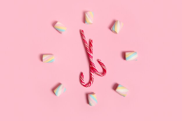 Top view with colorful sweet marshmallows and striped Christmas lollipops, candy canes stacked in the shape of a clock and dial, isolated on pink pastel colored background with copy space for ad