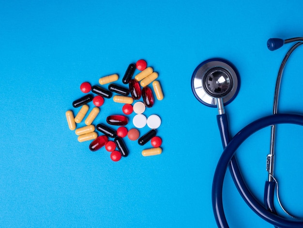 Top view with multicolored pills and capsules next to a stethoscope. Blue background and copy space
