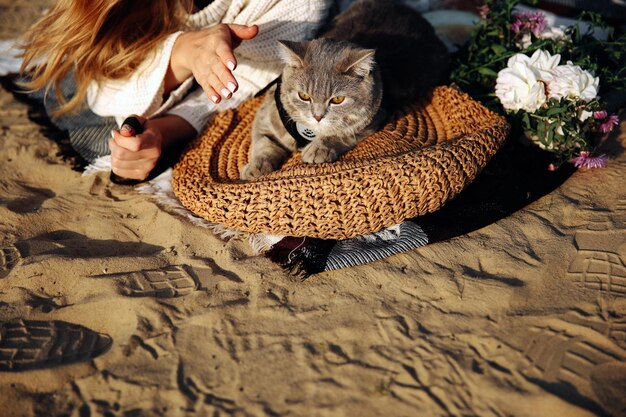 Top view of a woman on the beach with her kitten breed Scottish Rectangular gray at sunset The cat lies on a straw bag near the flowers