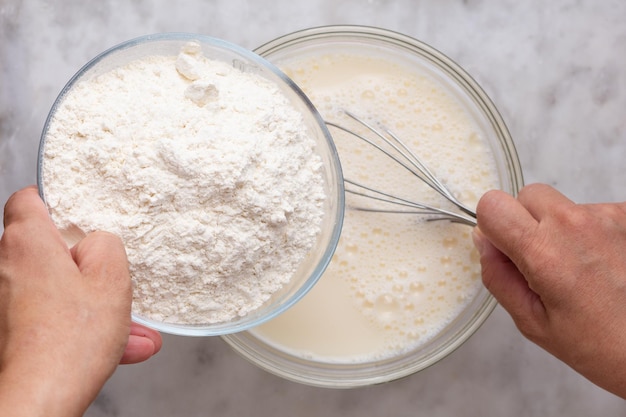 Top view of woman hand holding bowl with flour and putting into batter in glass bowl on the marble surface