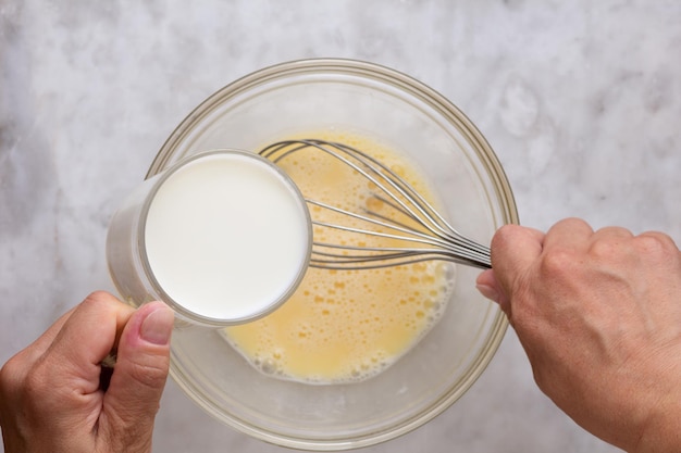 Top view of woman hand holding glass of milk putting into mixed raw eggs in glass bowl on the marble surface