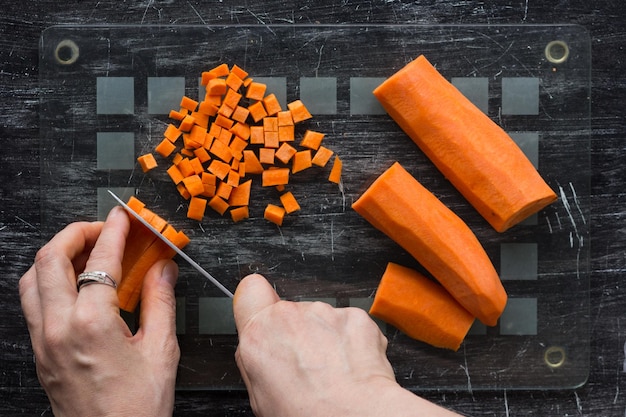 Вид сверху на женские руки, нарезающие сырую морковь на маленькие квадратные ломтики ножом на черном фоне
