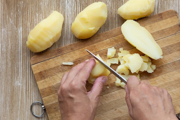 Foto vista dall'alto delle mani della donna che tagliano le patate bollite lavate in giacche usando il coltello sullo sfondo di legno