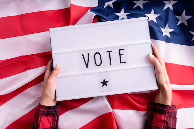 Photo top view of woman hands holding lightbox with the word vote on american flag background flat lay