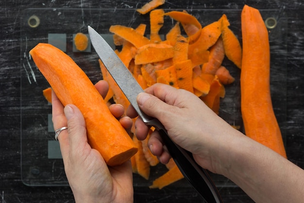 Vista dall'alto delle mani della donna che tengono la carota sbucciata e il coltello sul tagliere di vetro con peeling sullo sfondo nero