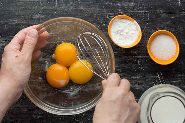 Top view of woman hands holding whisk and bowl with three yolks, starch, sugar and milk for mixing and making custard cream on the black background