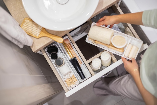 Vista dall'alto delle mani della donna che organizzano ordinatamente prodotti da bagno e articoli da toeletta nel cassetto del bagno