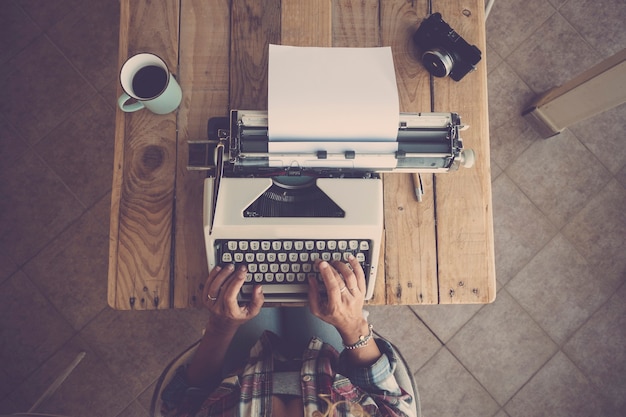 Foto vista dall'alto delle mani della donna che digitano sulla tastiera della macchina da scrivere su carta. donna che digita sulla tastiera della vecchia macchina da scrivere alla scrivania dell'ufficio con la tazza da caffè e la macchina fotografica sul tavolo