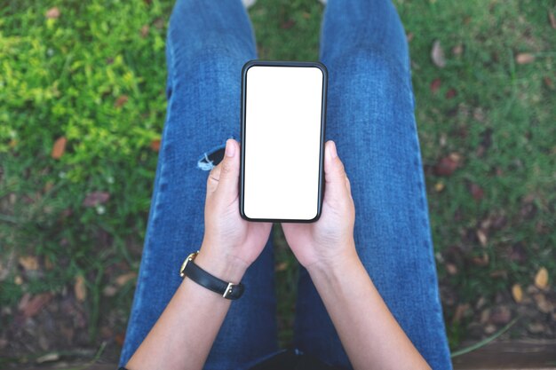 公園に座っている間空白の白い画面で黒い携帯電話を保持している上面図の女性