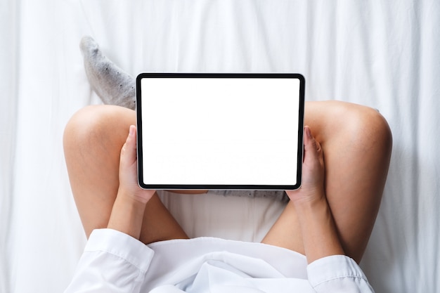 집에서 아늑한 흰색 침대에 앉아있는 동안 빈 바탕 화면 흰색 화면이 검은 태블릿 PC를 들고 여자의 상위 뷰