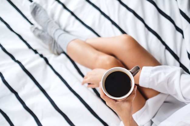 Top view  of a woman holding and drinking hot coffee in a white cozy bed at home in the morning