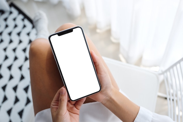 Vista superiore del telefono cellulare della tenuta della donna con lo schermo bianco da tavolino in bianco mentre sedendosi nella camera da letto con la sensibilità rilassata