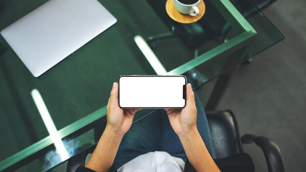 Вид сверху женщины, держащей мобильный телефон с пустым белым экраном рабочего стола с ноутбуком и чашкой кофе на столе