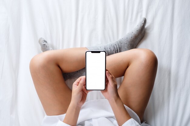 Punto di vista superiore di una donna che tiene telefono cellulare bianco con lo schermo da tavolino in bianco mentre trovandosi su un letto bianco accogliente a casa